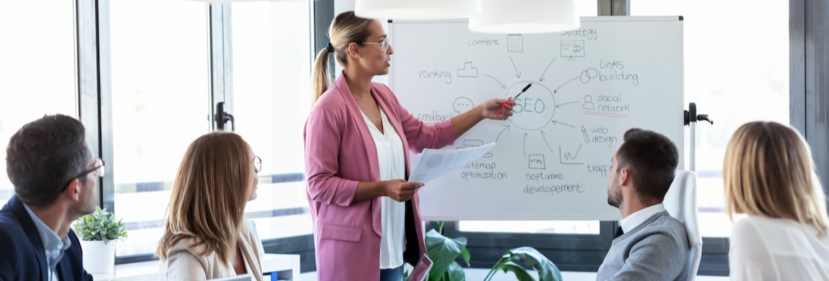 Elegant young businesswoman pointing at white blackboard and explain a project to her colleagues on coworking place.