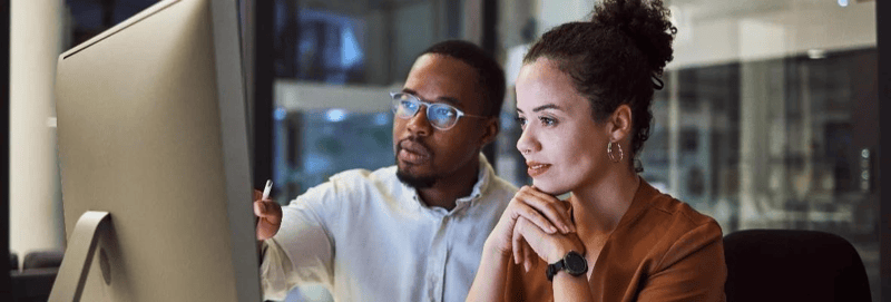 Two coworkers look at a computer screen and analyze data together. 