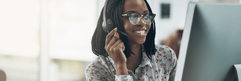 Black woman smiling while holding her headphone set and looking at a computer