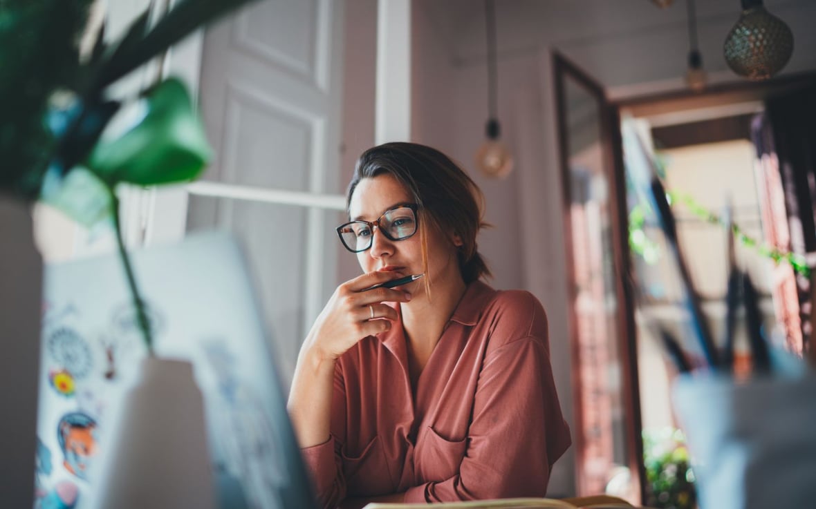 Caucasian woman with glasses holding a pen while looking at her laptop